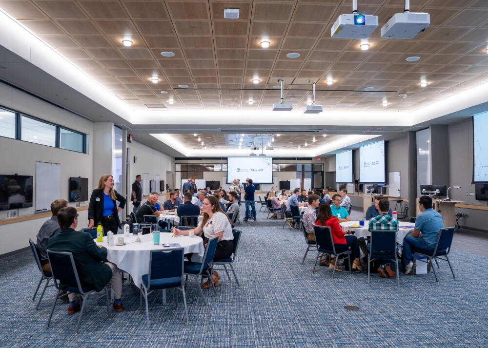 People seated in small groups at round tables during a professional event in a large, modern conference room with presentation screens and ceiling projectors.